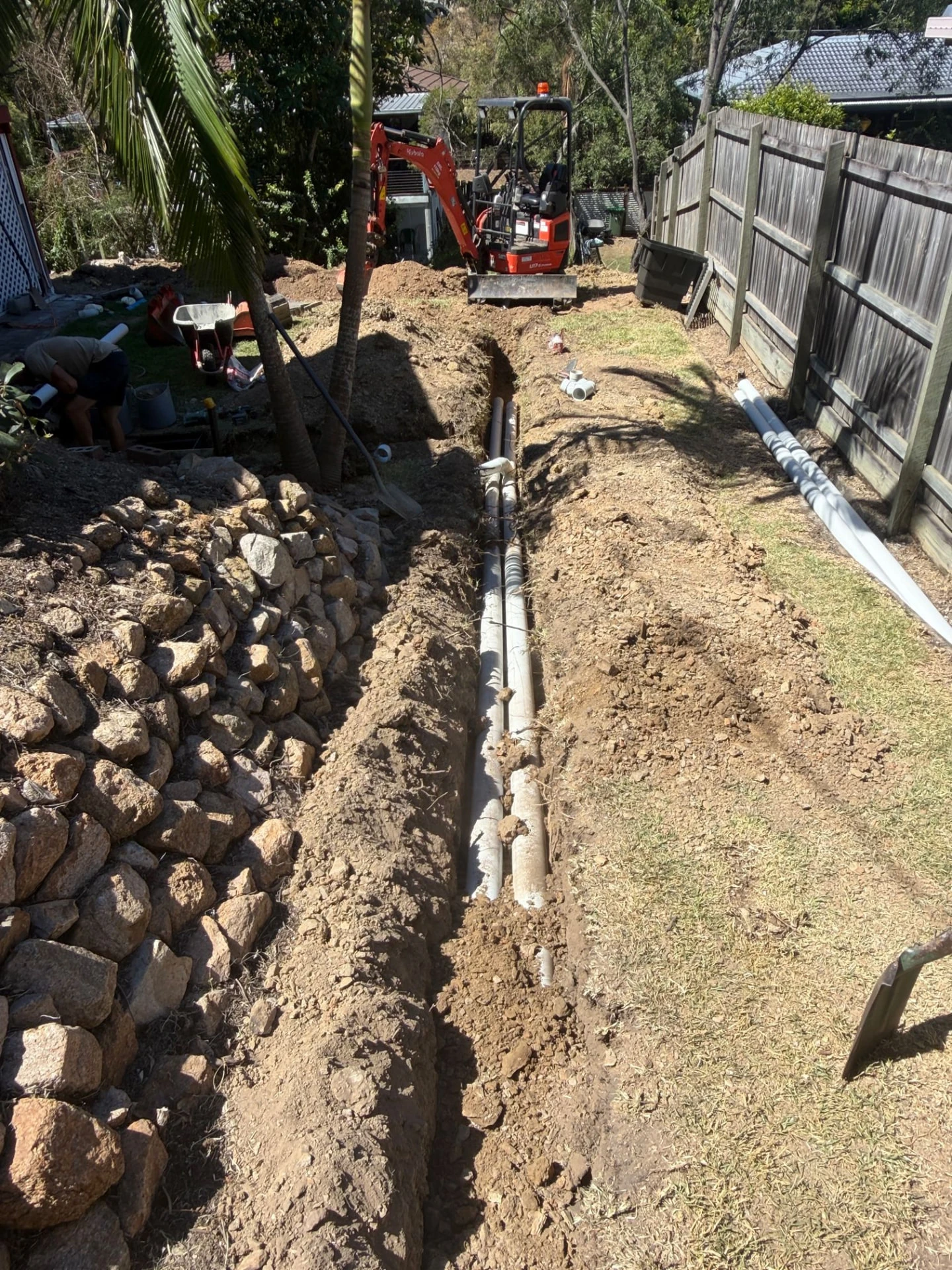 New drainage pipework laid in a Brisbane backyard trench with an excavator and spare PVC pipes on site