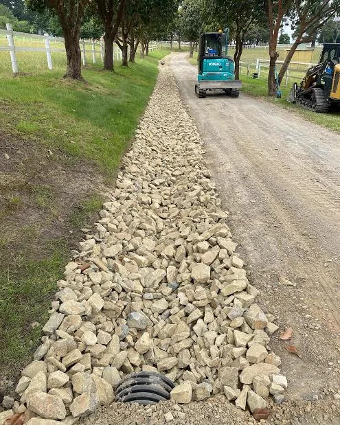 Rock-lined spoon drain beside a rural driveway in Brisbane with an excavator on site